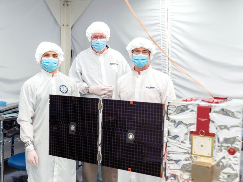 Gowned and masked engineers behind a satellite in a cleanroom.