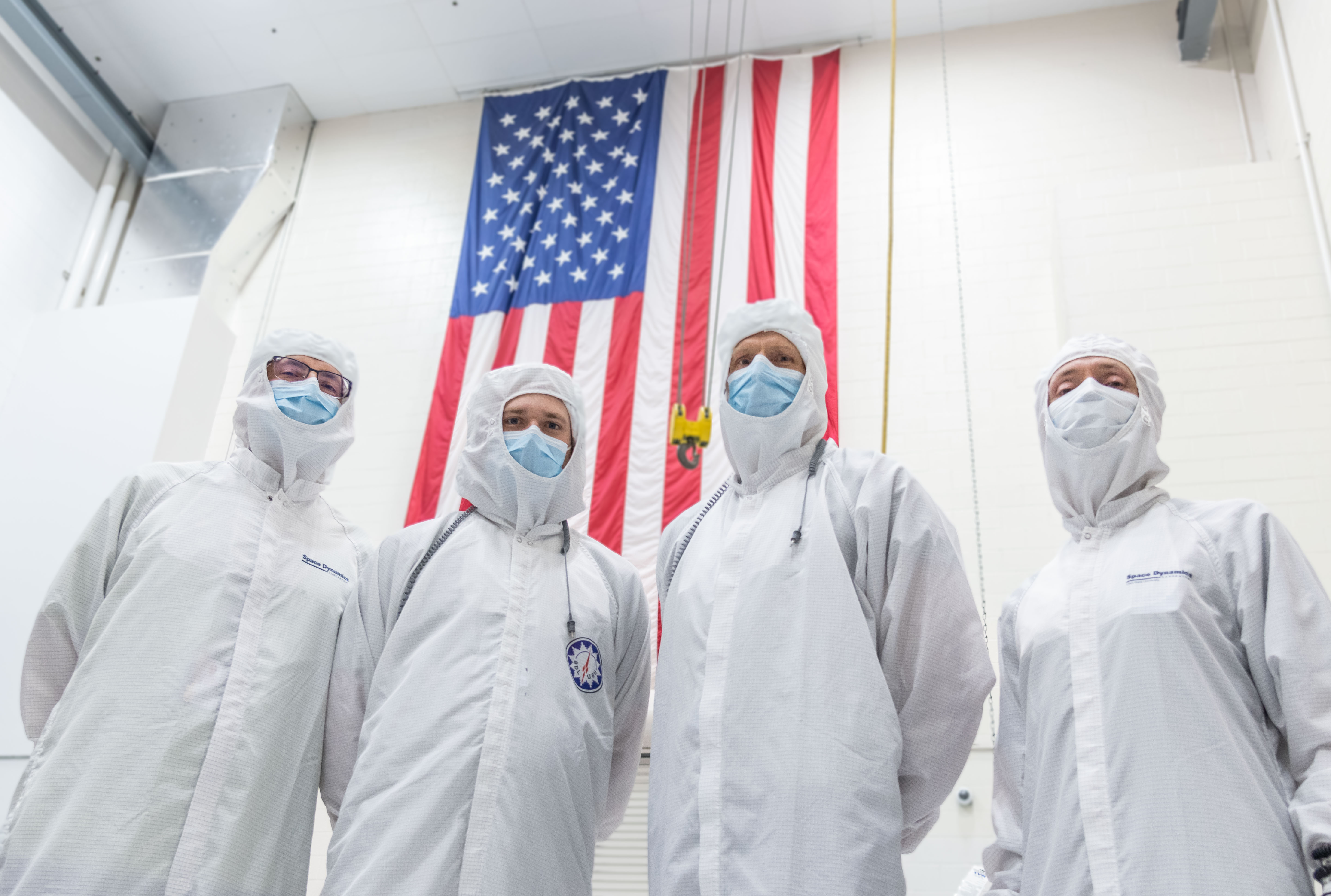 Engineers dressed in cleanroom gowns stand beneath the US flag.