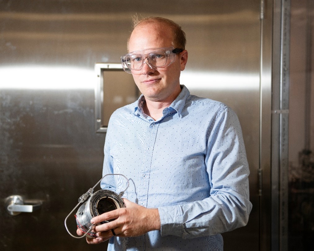 Tyson Smith standing inside a laboratory holding a piece of hardware.